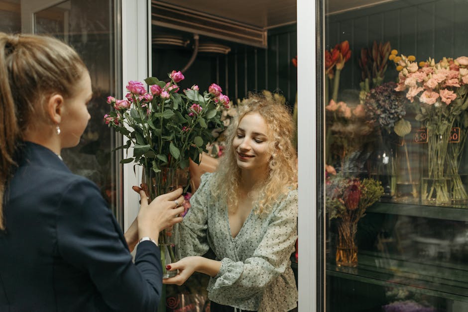 A cheerful woman with curly blonde hair and a patterned blouse is receiving a bouquet of fresh pink roses from a florist, a woman with brown hair tied back and wearing a navy blazer. The florist is ha