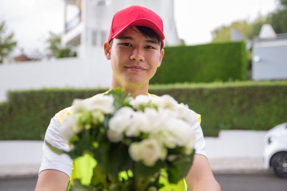 A young male flower delivery courier wearing a bright red cap and a neon yellow safety vest extends a bouquet of white flowers towards the camera, with a friendly expression in an outdoor residential