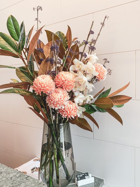 A tall, clear glass vase filled with a fresh floral arrangement featuring soft pink chrysanthemums, delicate white orchids, and sprigs of purple lavender. The bouquet is complemented by greenery with