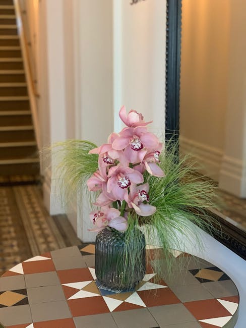 A tall, elegant floral arrangement in a dark, ribbed glass vase situated on a decorative tiled table. The bouquet features soft pink orchids with multiple blossoms cascading along green stems, complem