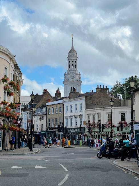 The image depicts a lively street scene in a historic town with a prominent clock tower featuring a golden weather vane at the top, set against a partly cloudy sky. Surrounding the tower are tradition