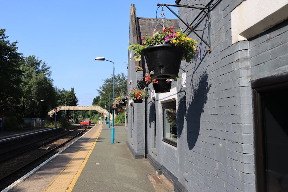 A row of black hanging flower baskets containing colorful blooms, including pink, yellow, and purple flowers, is suspended from wrought iron brackets on the side of a gray brick building at Motspur Pa