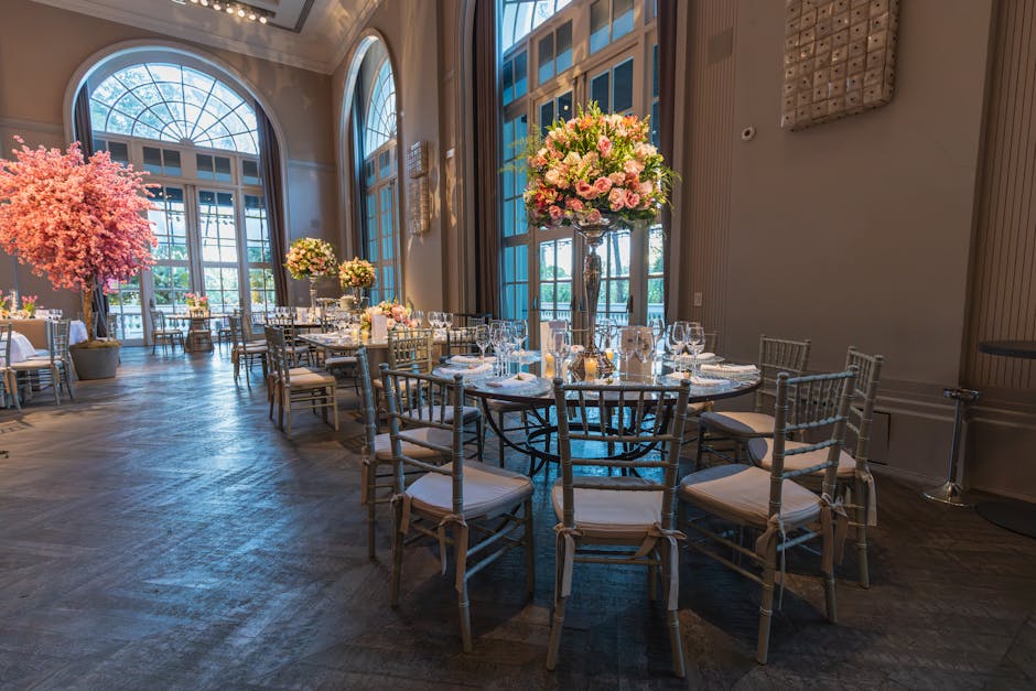 An elegant banquet hall set up for a wedding reception featuring a central round table decorated with a large floral arrangement of pink and white roses, peonies, and greenery placed in a tall glass v