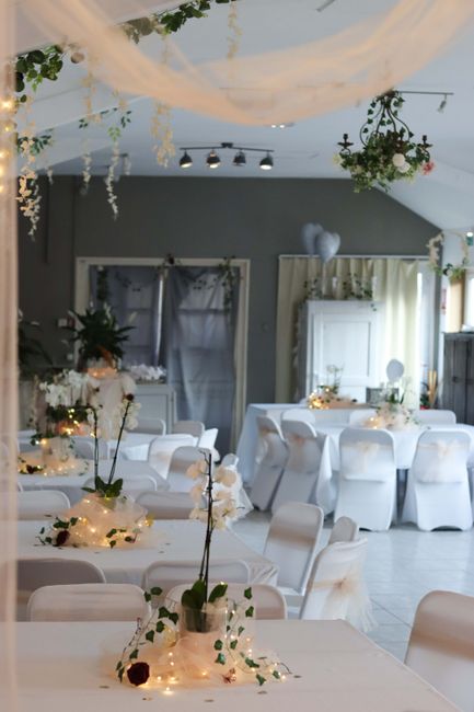 The image showcases an elegantly arranged interior space set up for a wedding or special event, featuring tables decorated with floral arrangements and soft lighting. In the foreground, a table is ado