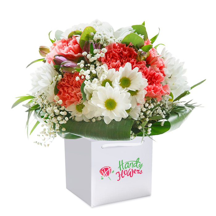 Upright and elegantly poised, a freshly assembled bouquet sits grandly in a clean, square white gift bag, with soft light casting gentle shadows on its radiant contents. Jade-green foliage forms a deep, glossy backdrop, creating a natural embrace for the vivid coral and peach carnations whose abundant, ruffled petals bring lively texture. Amongst them, snowy white daisies with striking lime-green centers beam like little sunbursts, while billowing sprigs of baby's breath provide a subtle frothiness throughout. Look closer and you'll spot the subtle intrigue of half-hidden, purple-tinged buds tucked thoughtfully on the left side-perhaps a nod to the blooming trees along Alexandra Avenue. All flowers are arranged in a lush, dome-like shape, rich with colour and freshly-cut vibrance. The balanced composition is enhanced by a crisp white gift bag, its ribbon handles elegantly knotted, designed for effortless handover at a front door or picked up on the way to a gathering near Motspur Park station. This bouquet captures the spirit of the local community: inviting, cheerful, and ready to mark celebrations both big and small. A perfect example of the artistry and care you'd expect from your friendly Motspur Park florist, this arrangement promises to create smiles and lasting memories.